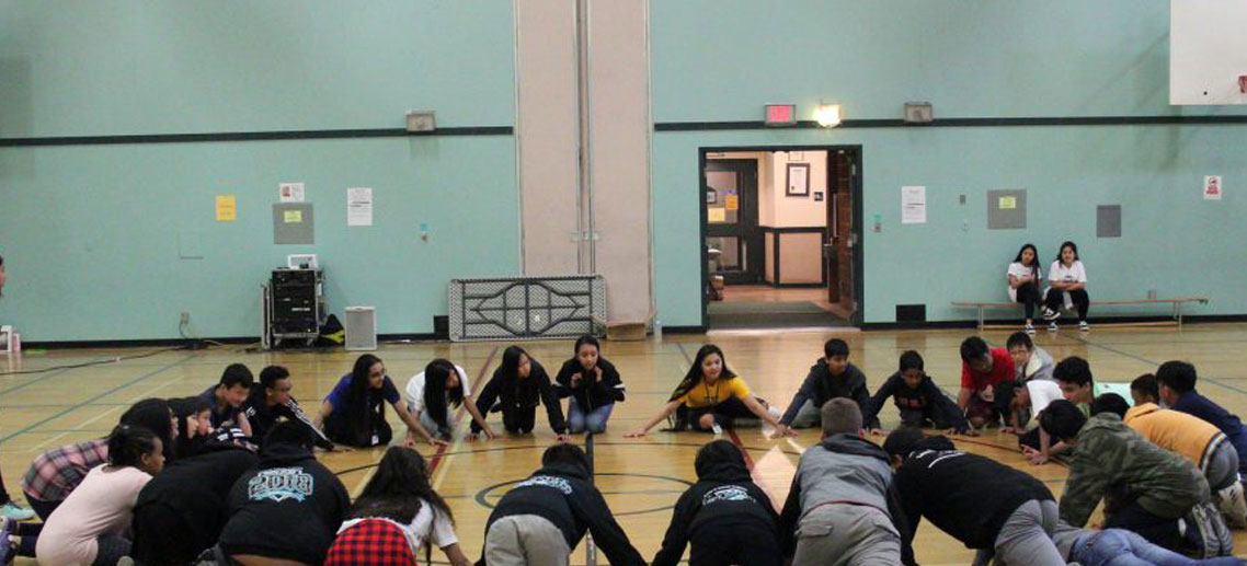 a large group of children in a gymnasium, sitting in a circle, about to play a game
