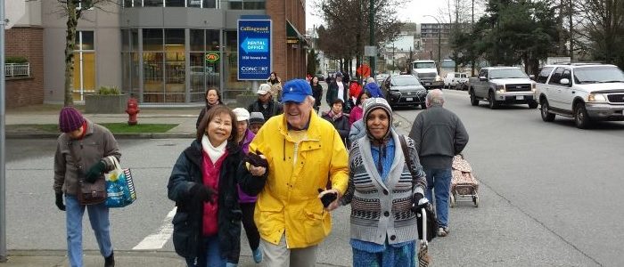 a group of seniors walking together on Joyce Street and smiling. Two seniors hold the hands of a blind man and help him to walk