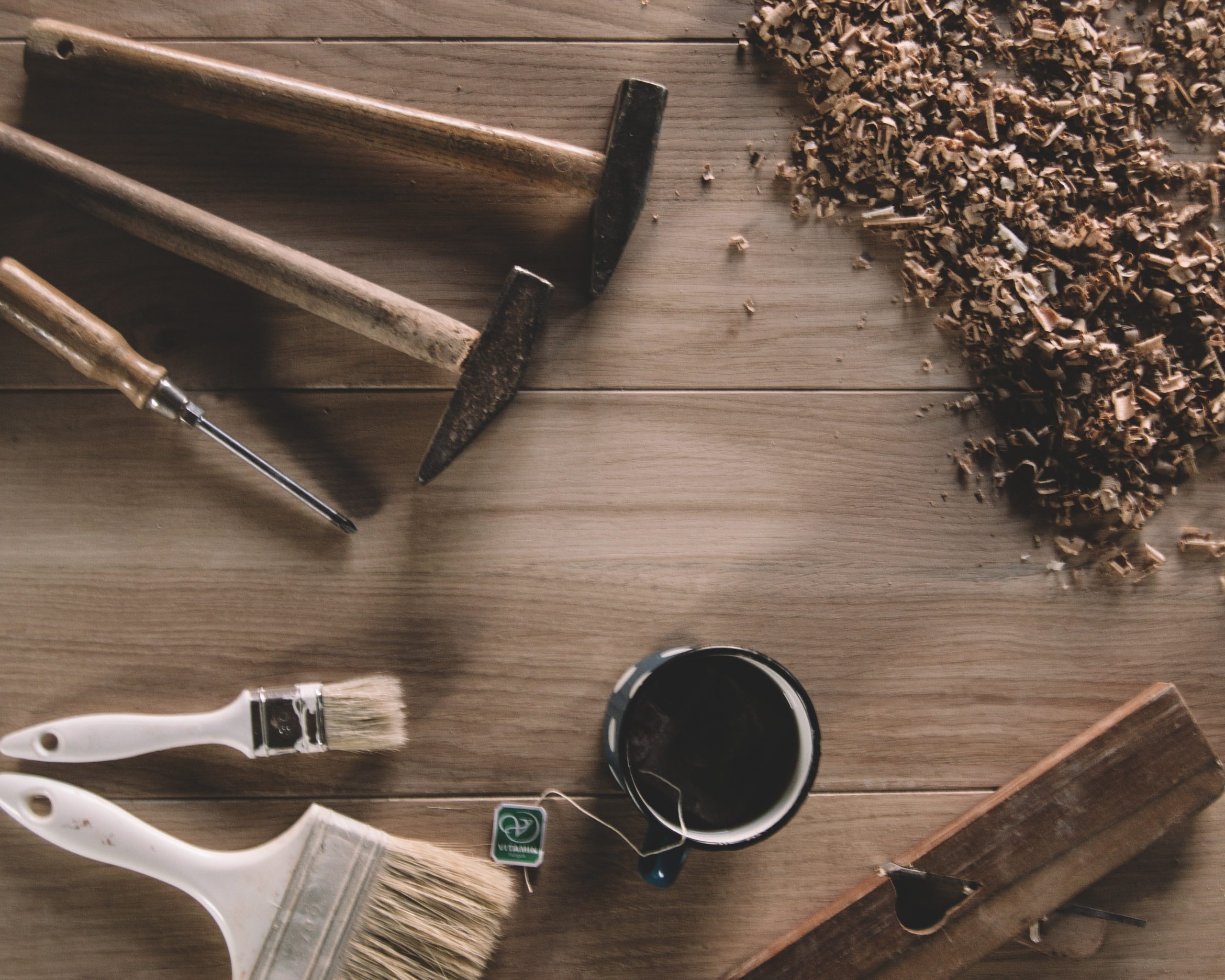 Tools, paintbrushes and wood shavings are arranged on a wooden table. There is a cup of tea next to the tools.