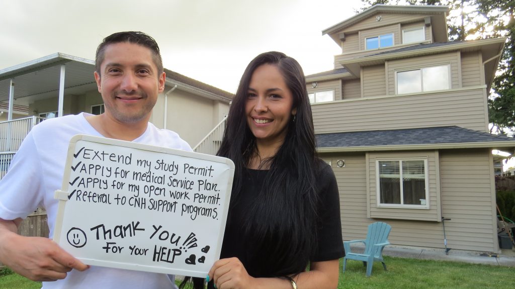 Rafael and Yudy Kainan stand outside holding a whiteboard. It reads, checked off, "extend my study permit, apply for medical service plan, apply for my open work permit, referral to CNH support programs. Below, it reads, "thank you for your help" with a smiley face and hearts.