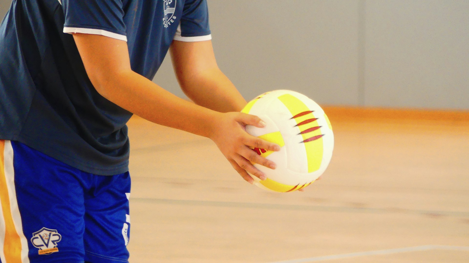 A pair of hands holds a volleyball inside a gym
