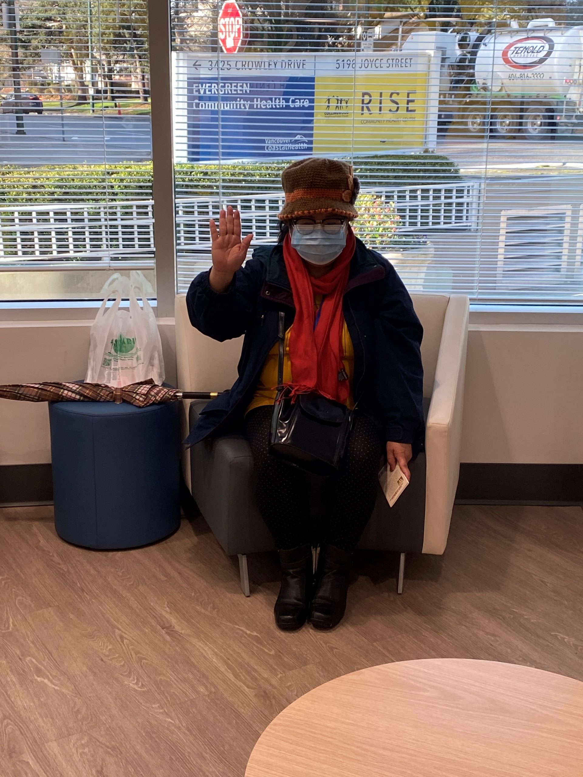 An older woman sits on a chair in a lobby, waving towards the camera. She is wearing a dark jacket, red scarf, toque, and face mask.