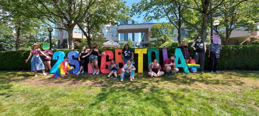 A group of teenagers sit and stand outside in front of large, colourful letters that read 2SLGBTQIA+