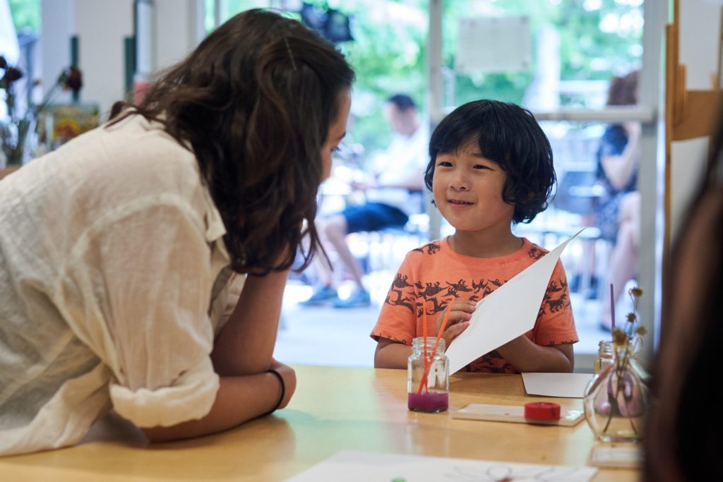 A child wearing an orange t-shirt holds a piece of paper while smiling towards an adult