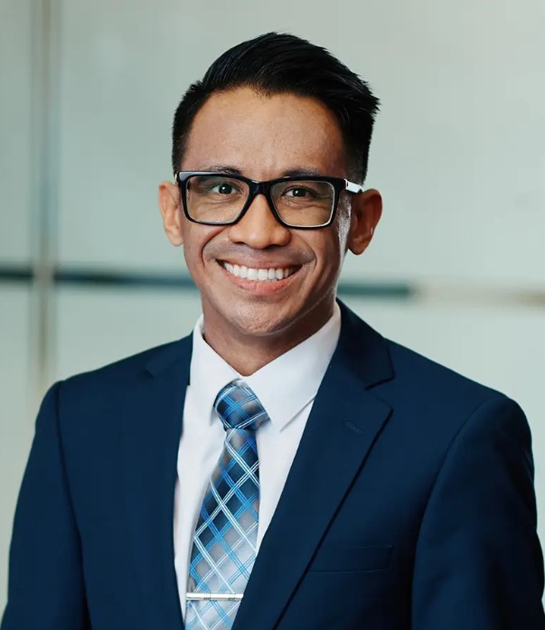 A close-up of a man wearing glasses and a dark blazer with white shirt and blue tie smiling towards the camera