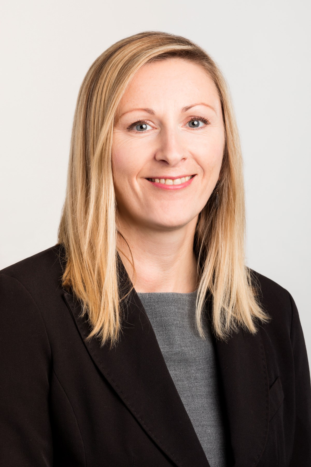 A close-up photo of a woman with shoulder-length blonde hair wearing a black jacket and grey shirt