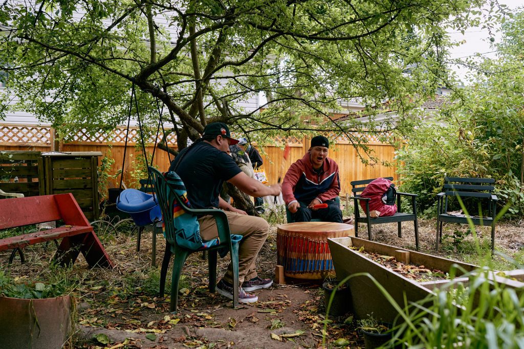 Two teenagers sit in a garden around a large drum on the ground. Together, they play the drum with mallets while singing.