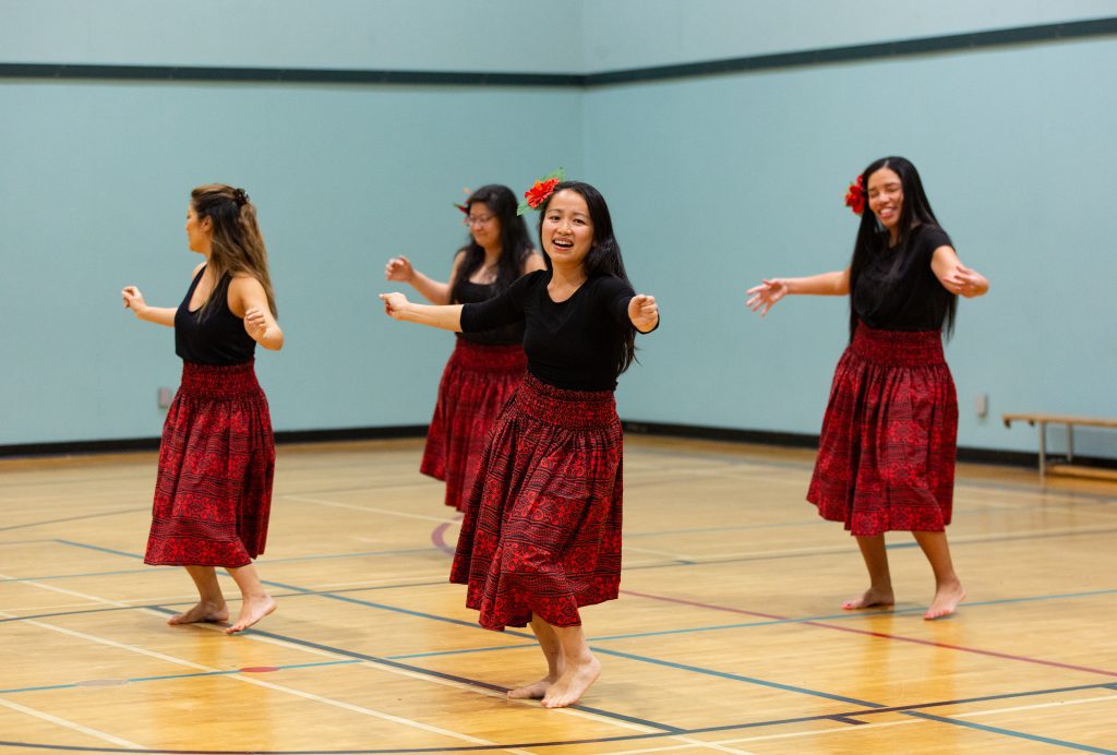 A group of four people with long hair dance bareboot wearing long red skirts and black shirts inside the gym at CNH