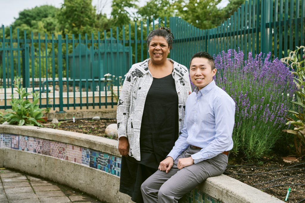 Two people pose for a photo on the rooftop garden at CNH. One is sitting while wearing a dress shirt and pants, while the other is standing wearing a long black dress and shirt patterned with newsprint