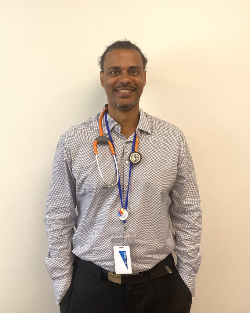Clement Bélanger Bishinga stands against a plain white wall at RISE Community Health Centre. Smiling with his hands in his pant pockets, he is wearing an orange stethoscope and blue lanyard around his neck.