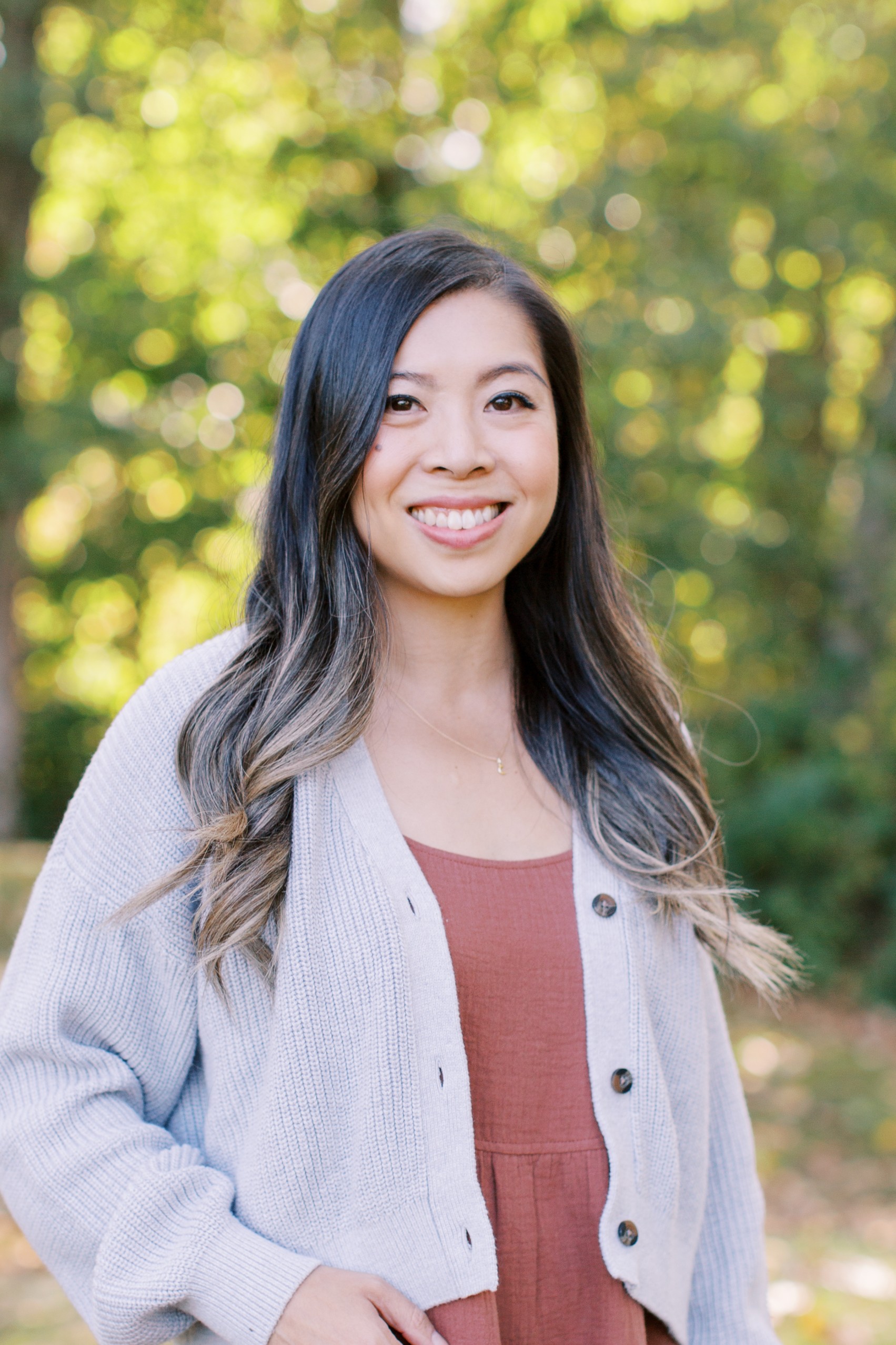 A close-up of a woman with long dark hair wearing a light-coloured cardigan stands outside, smiling towards the camera