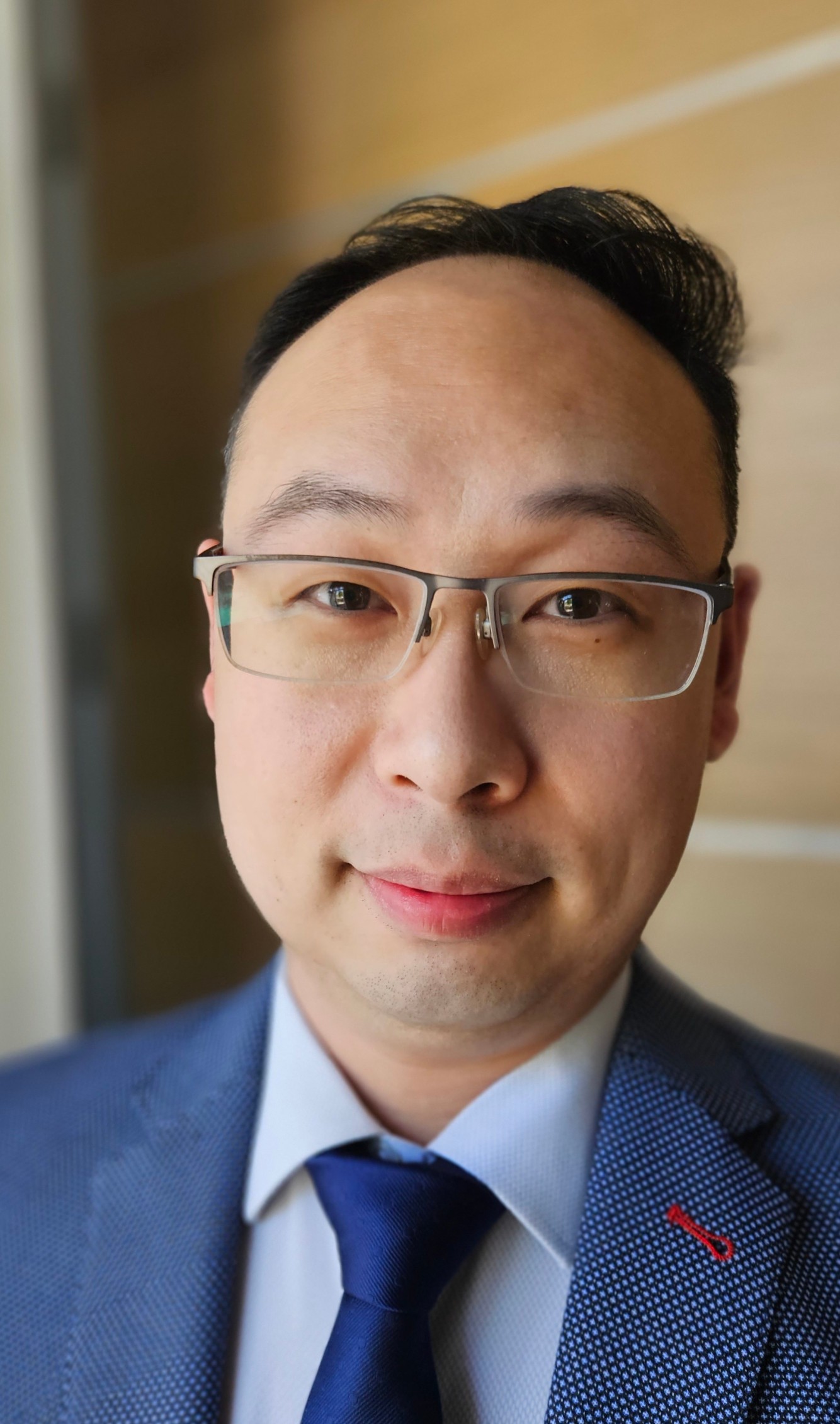 A close-up of a man with short dark hair wearing glasses, a blue blazer and tie smiling towards the camera