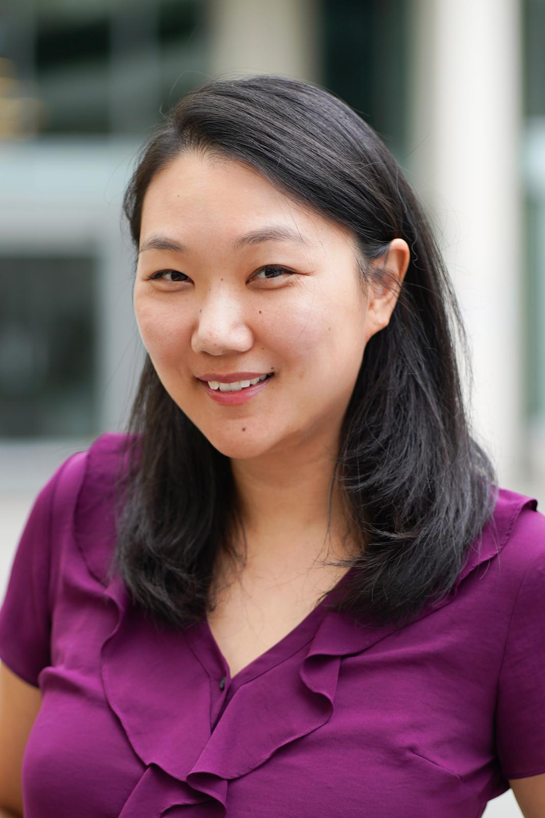 A close-up of a woman with dark shoulder-length hair wearing a purple blouse smiling towards the camera