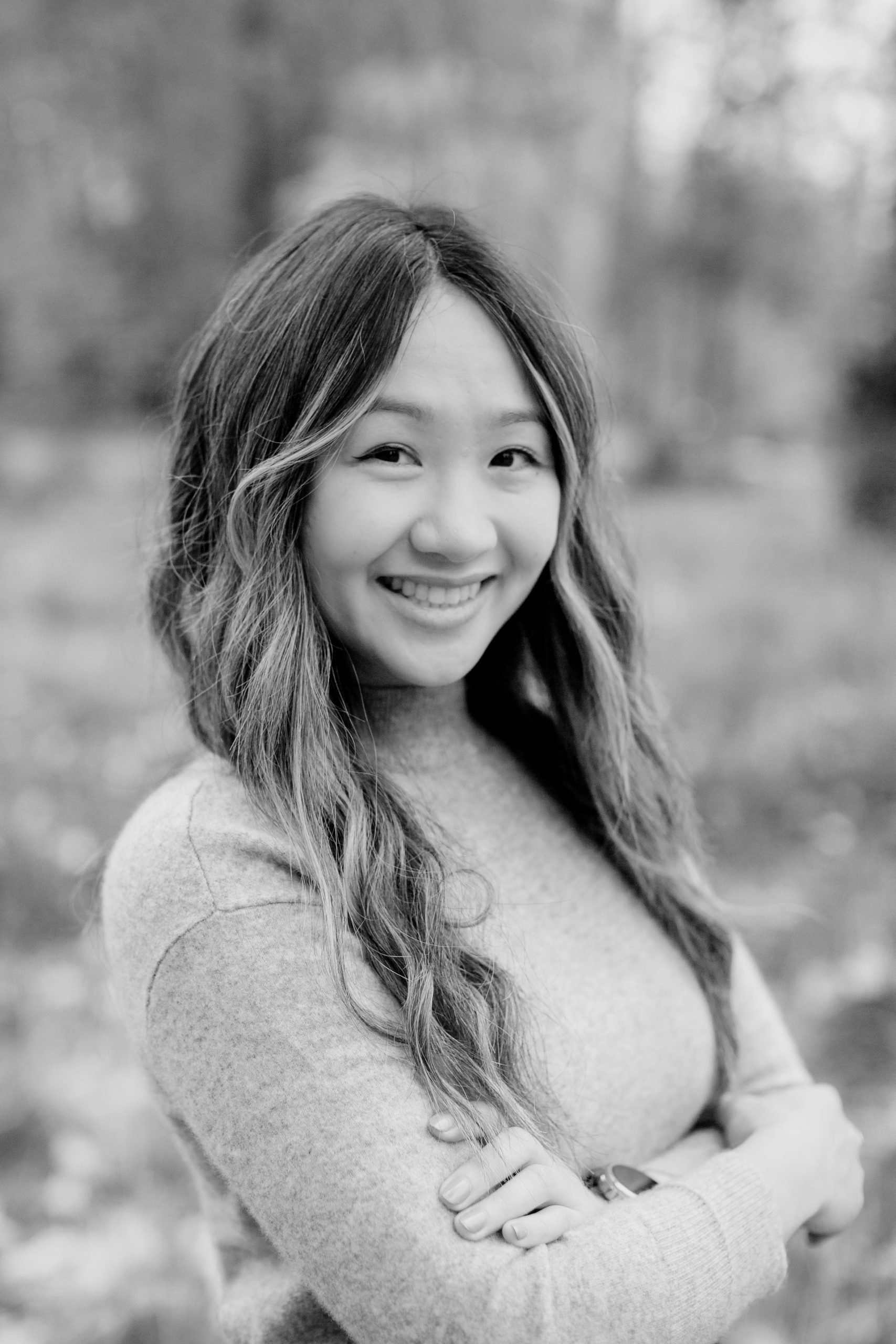 A close-up of a woman with long curly hair standing outside with her arms folded. She is smiling towards the camera.