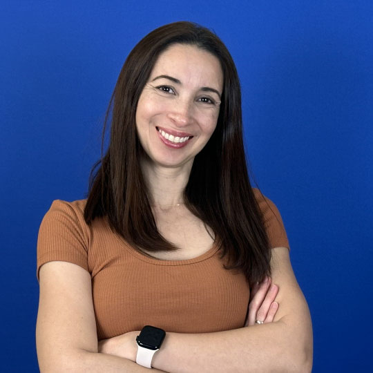 A photo of Liz Ayda Ramirez standing with her arms folded and smiling towards the camera against a blue background
