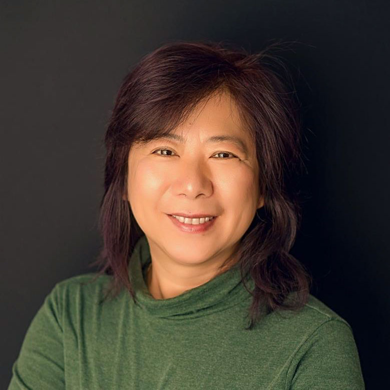 A close-up of a woman with shoulder-length brown hair and side swept bangs smiling towards the camera. She is wearing a green turtleneck.