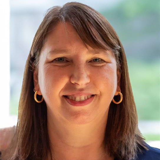 A close-up of a woman with shoulder-length brown hair and side bangs smiling towards the camera.