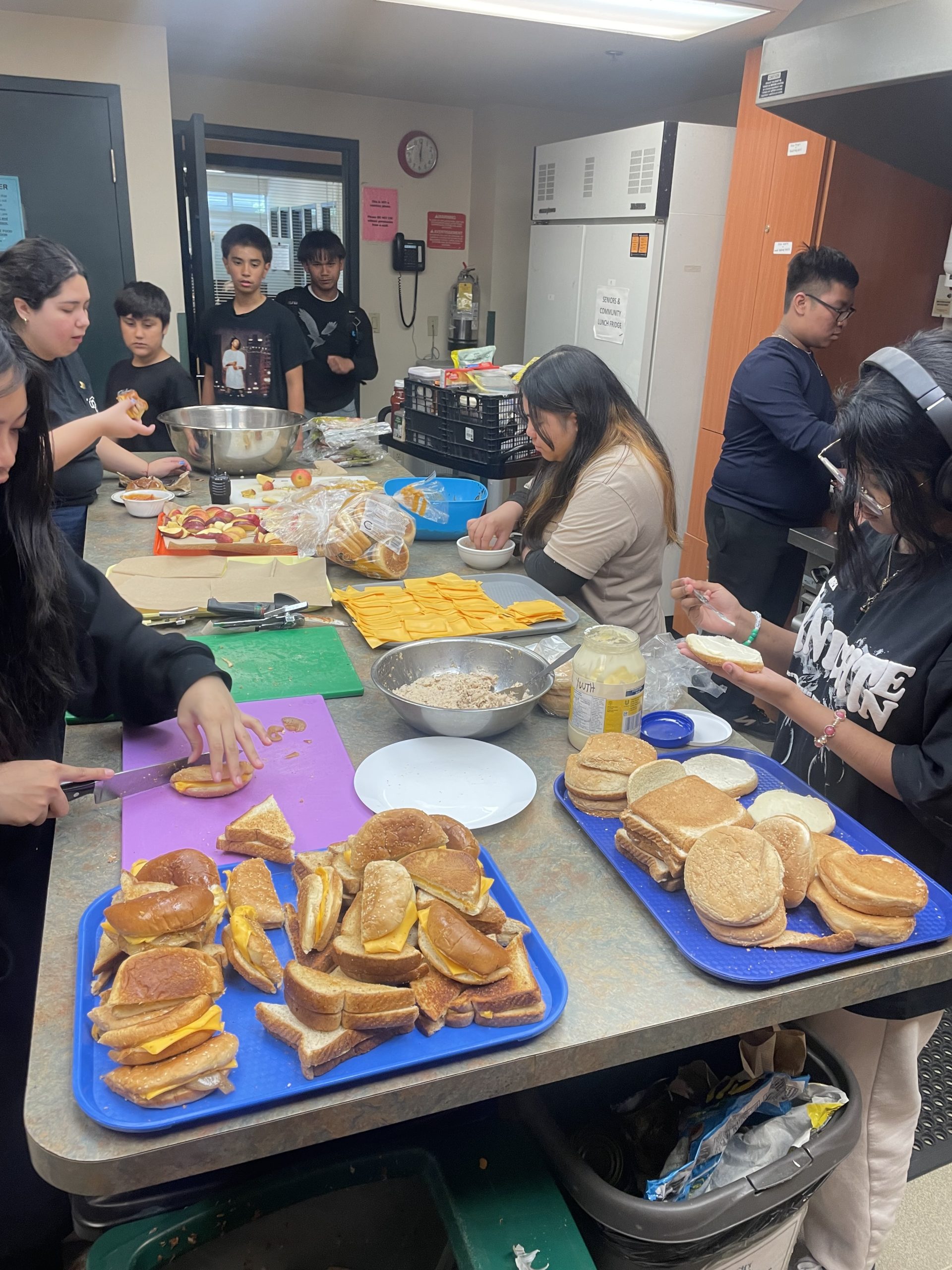 A group of teenagers stand inside a commercial kitchen making sandwiches