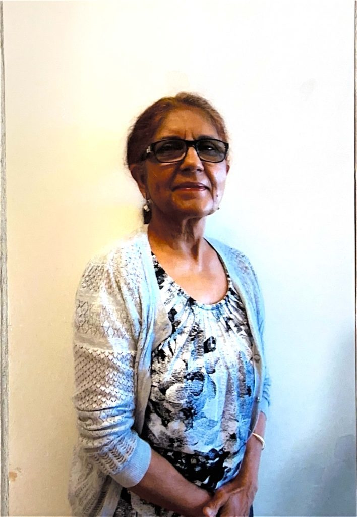 A woman with brown hair pulled back into ponytail. She is wearing glasses and a grey cardigan over floral blouse.