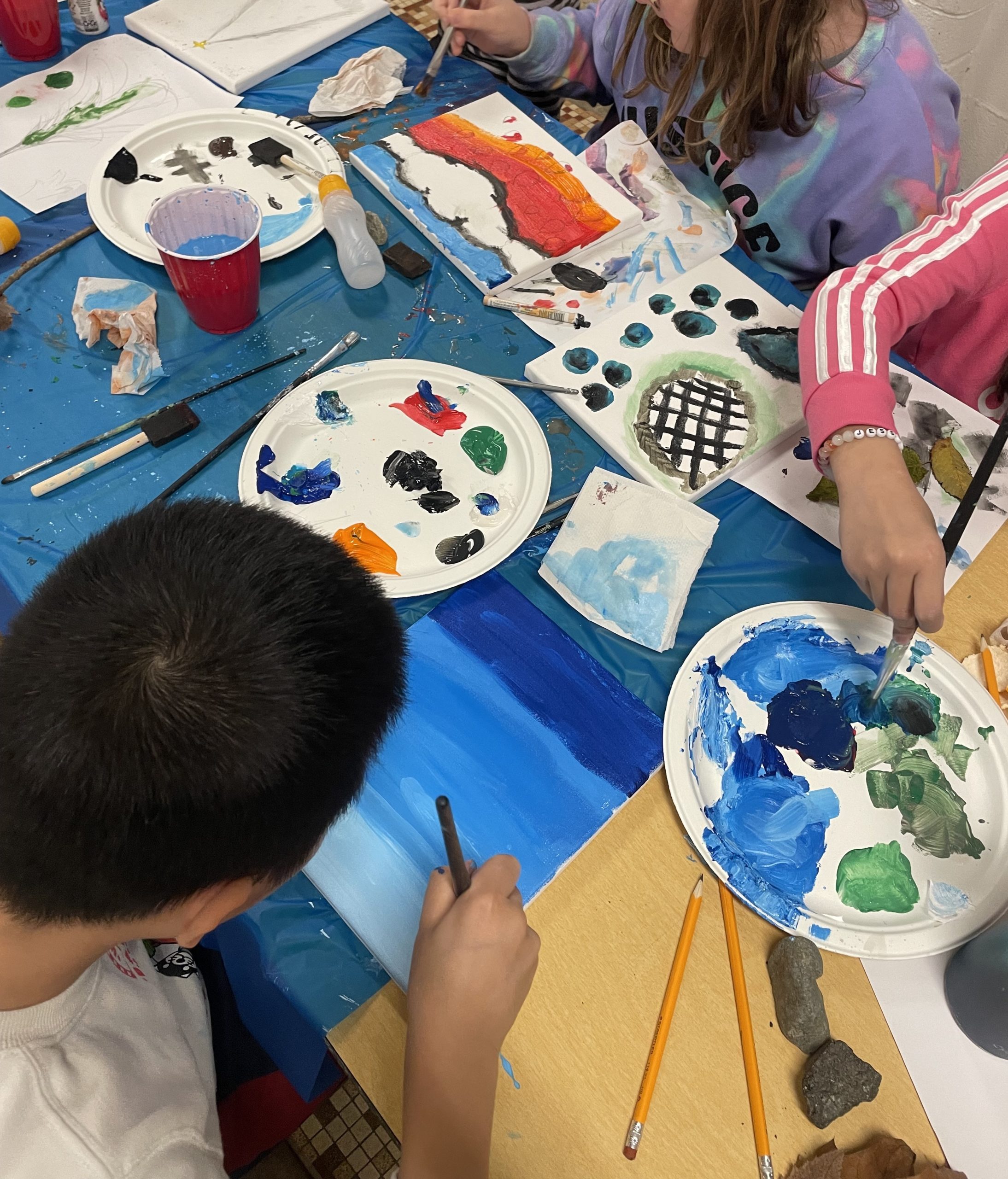 Paint palettes, brushes and paper are sprawled onto a table with two teenagers hunched over the table painting