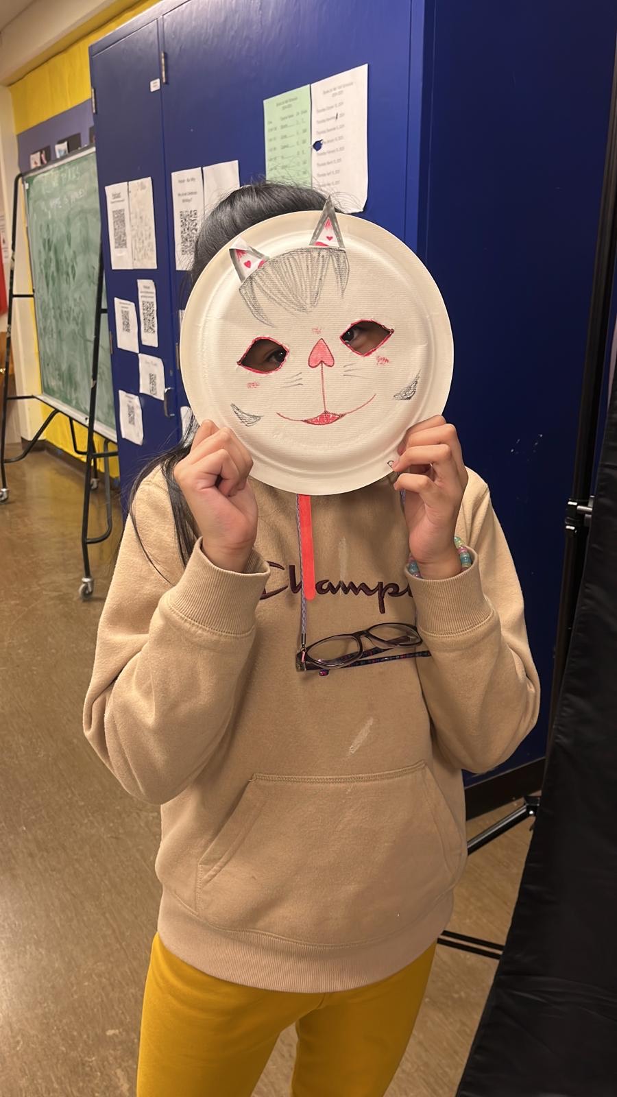 A teenager wearing a tan sweater holds a paper plate over their face. The plate has two holes cut out for the eyes and drawings to illustrate a cat's face.