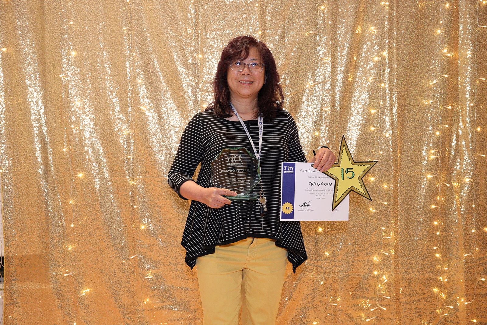 A woman stands in front of a golden backdrop holding a paper certificate and a yellow star that says, "15"