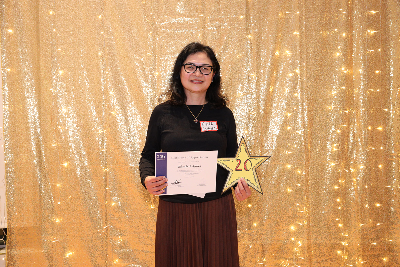 A woman stands in front of a golden backdrop holding a paper certificate and a yellow star that says, "20"