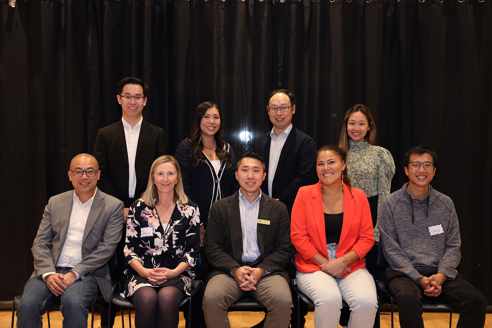 A group of nine people sitting and standing in two rows in front of a black curtain. They are smiling towards the camera.