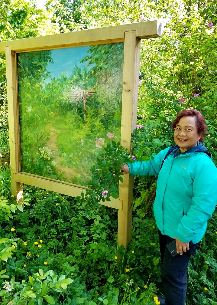 A woman wearing a teal jacket stands next to a large painting standing inside a garden