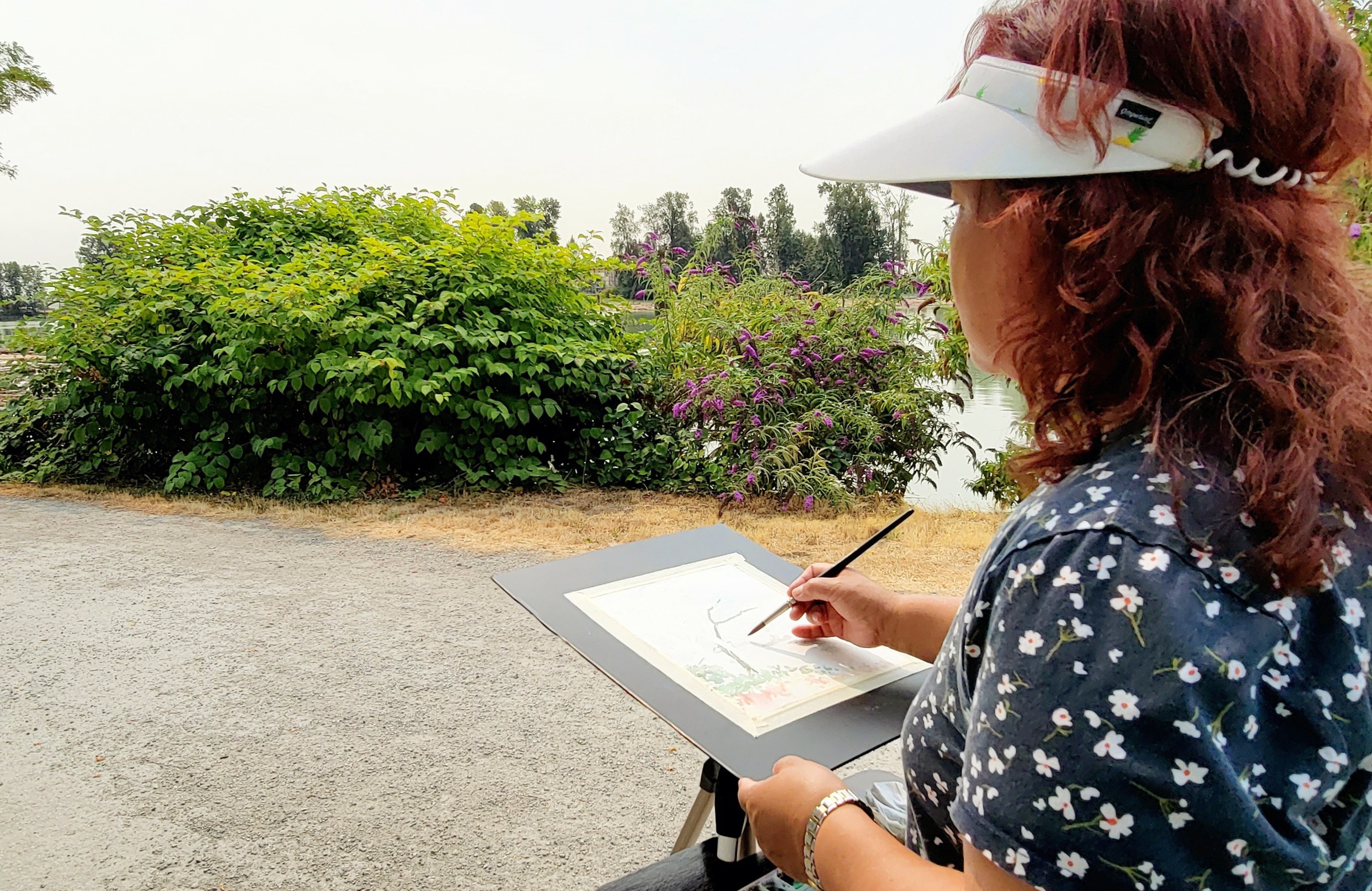 A woman with curly red hair sits outside painting a scene along the Fraser River