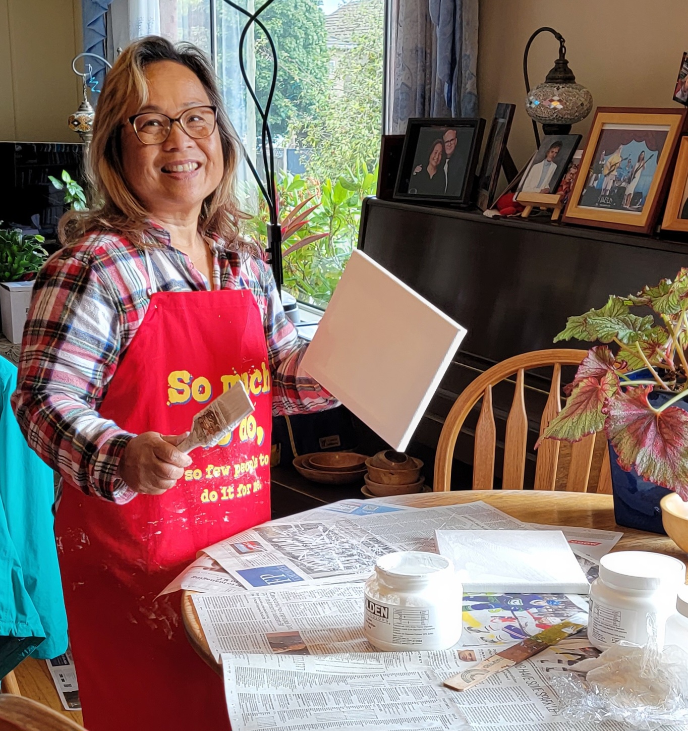 A woman wearing an apron stands next to a table holding a blank canvas and a paintbrush