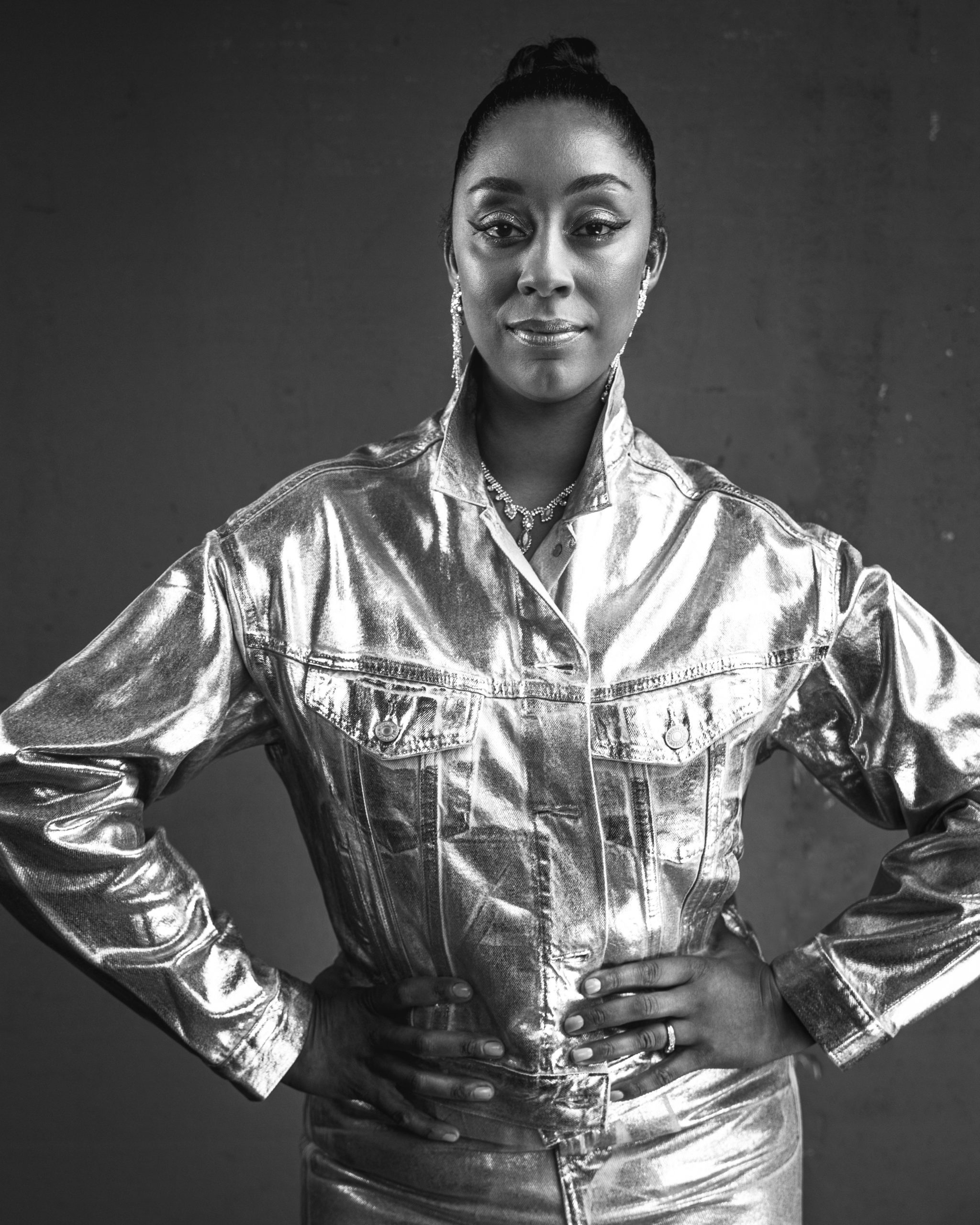 A black and white photo of Krystle Dos Santos standing in a metallic jacket with her hands on her hips
