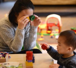 A mother holding a triangle and circle toy block with holes against her eyes and looking through at a child, holding a pentagon block.